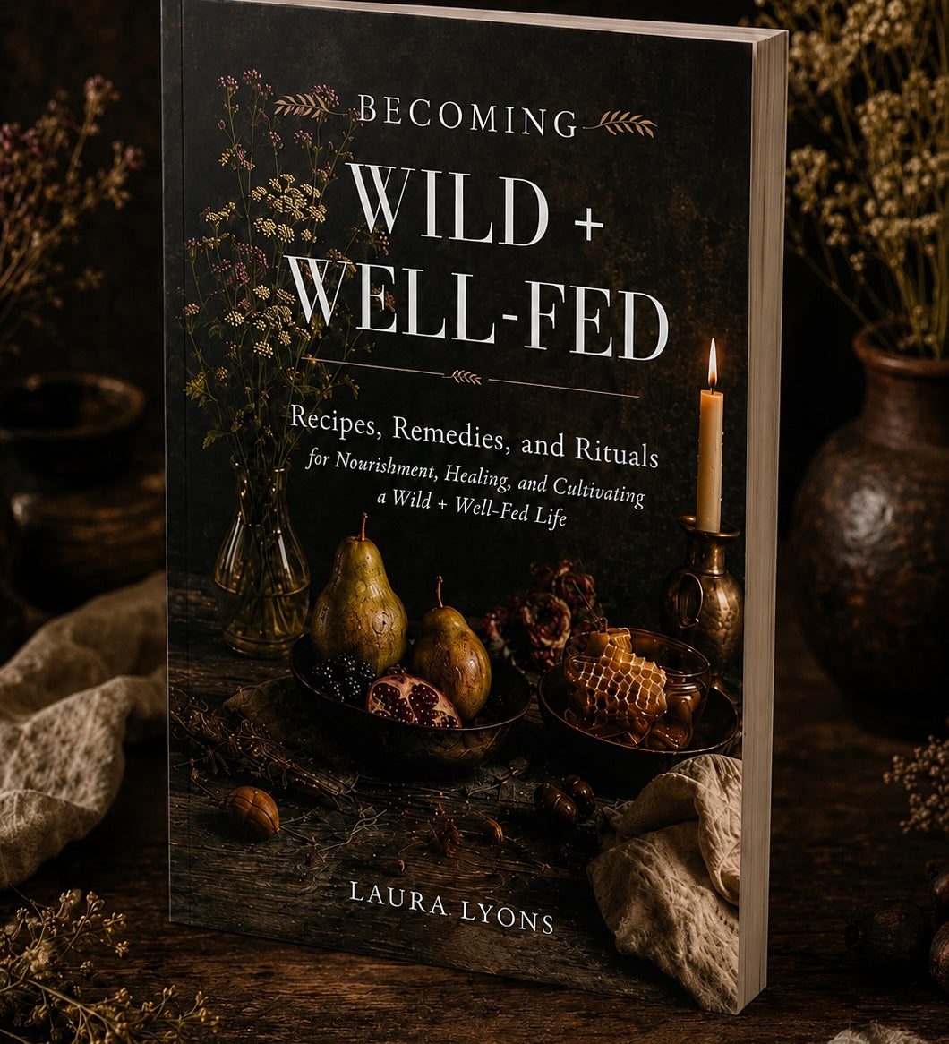 A softcover book titled 'Becoming Wild + Well-Fed' by Laura Lyons, standing upright on a rustic wooden surface with dried flowers and candles in the background.