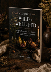 A softcover book titled 'Becoming Wild + Well-Fed' by Laura Lyons, standing upright on a rustic wooden surface with dried flowers and candles in the background.