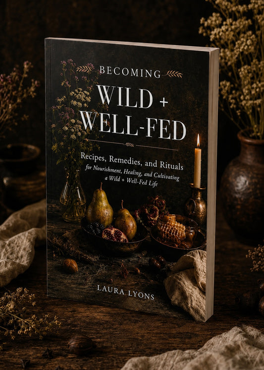A softcover book titled 'Becoming Wild + Well-Fed' by Laura Lyons, standing upright on a rustic wooden surface with dried flowers and candles in the background.